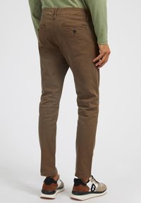 Pantalon en coton marron à coupe slim, doté de poches arrière et d'une petite étiquette de marque. Porté avec des baskets multicolores.
