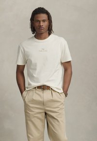En ung man med korta dreadlocks som bär en vit Polo Ralph Lauren t-shirt, beige veckade byxor och ett brunt bälte, står med händerna i fickorna.
