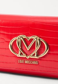Borsa in pelle sintetica rossa effetto coccodrillo con logo "M" dorato e testo "LOVE MOSCHINO" nella parte inferiore. Texture liscia e lucida.