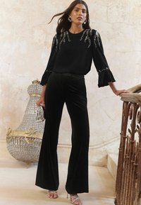 Top a maniche lunghe nera con decorazioni in paillettes, abbinato a pantaloni larghi neri in velluto; modella che tiene una pochette argentata e indossa tacchi argento.