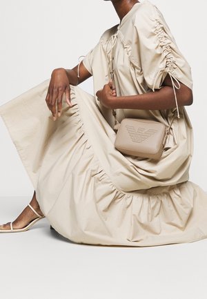 Béžová crossbody taška vyrobená z texturované kůže; obsahuje embosované logo a nastavitelné ramenní popruhy. Nositelná s volně střiženými béžovými šaty se staženými detaily.