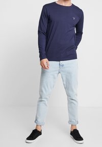 Maglietta a maniche lunghe blu navy con scollatura a girocollo e piccolo logo sul petto, abbinata a jeans chiari e scarpe da ginnastica nere.