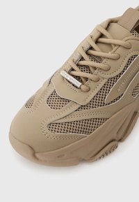 Béžové tenisky s mesh panely, šněrováním a metalickým logem "Steve Madden" na boku, které ukazují stylový sportovní design.