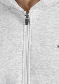 Detailní záběr na světle šedou mikinu na zip s viditelným zipem a malou částí krku osoby nad límcem.