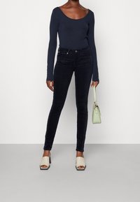 Mörkblå långärmad topp, mörka slim-fit jeans, beige klackskor med blockklack, håller en ljusgrön handväska; slät tyg och figurnära design.