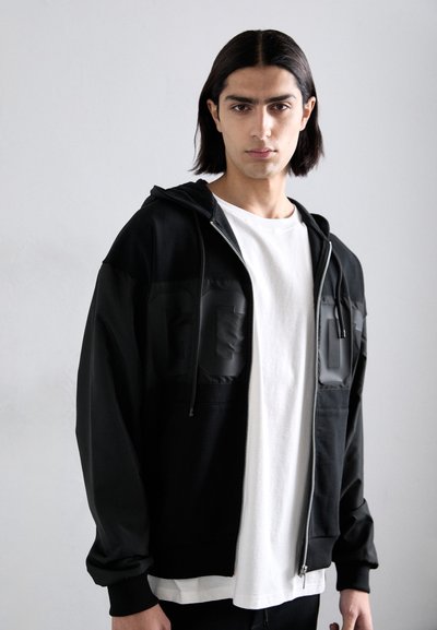 Schwarze Zip-Hoodie mit einer glänzenden Panele, die geprägte Buchstaben zeigt, aus leichtem Stoff und in lockerer Passform, kombiniert mit einem weißen T-Shirt.