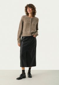 Bruine gebreide cardigan met knoopsluiting, gecombineerd met een zwarte leren midi rok. Model draagt zwarte kanten sokken en veterschoenen.