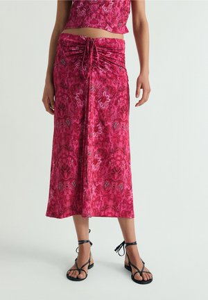 Mujer con una falda midi rosa floral con un lazo fruncido en la cintura y sandalias negras de tiras, de pie sobre un fondo liso.