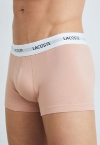 Lacoste férfi boxeralsó világos rózsaszín pamutból, széles fehér derékpánttal, fekete logóval, sima textúrával és alakra szabott fazonnal.
