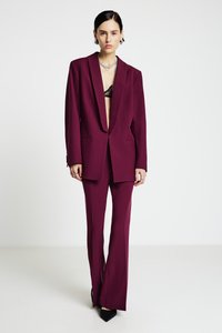 Costume sur mesure bordeaux comprenant un blazer croisé et un pantalon évasé, confectionné dans un tissu texturé. Les accents incluent des chaussures noires et un haut en dentelle.