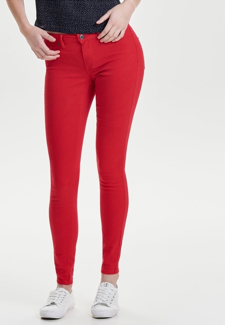 Jean skinny rouge à texture lisse, doté d'une fermeture à bouton et glissière à l'avant, associé à des baskets blanches et un haut bleu marine à pois.