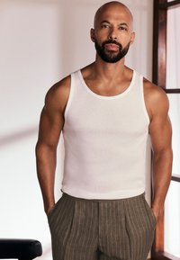 Débardeur blanc côtelé, pantalon beige à fines rayures, silhouette musclée, cheveux courts et barbe, debout devant un fond neutre.