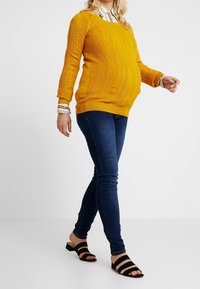 Keltainen neulottu äitiysneule, jossa on ribbi-tekstuuri ja palmikkoneule design, yhdistettynä tummansinisiin skinny farkkuihin ja mustiin hihnallinen sandaalit.