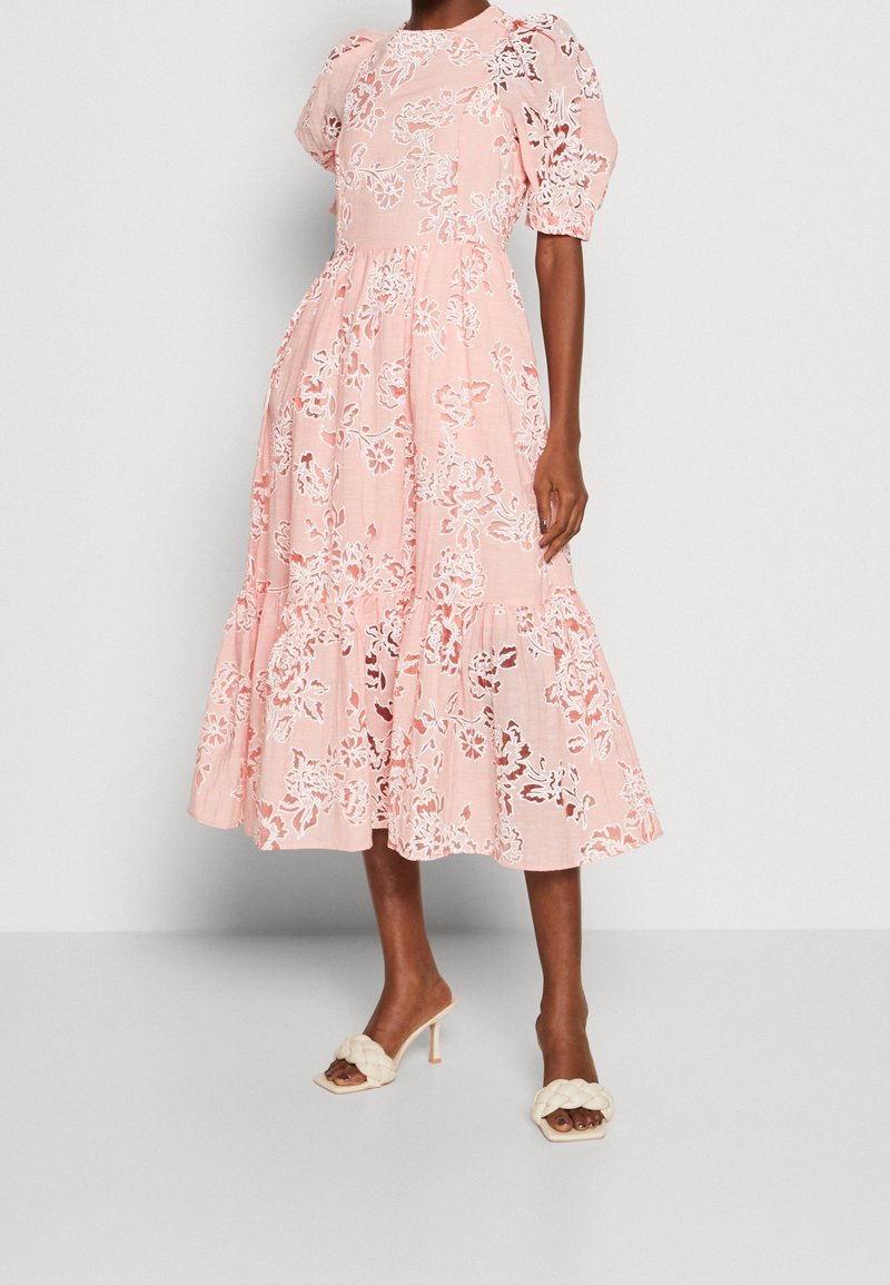 Femme portant une robe midi rose avec des détails en dentelle florale blanche et des manches bouffantes, associée à des sandales à talons hauts crème à brides tressées.