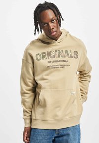 Beige hooded sweatshirt featuring a front pocket, bold "ORIGINALS" text, and ribbed cuffs, made of soft, textured fabric.