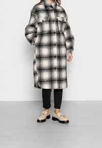 Lang ternet frakke med sorte, hvide og beige nuancer, to brystlommer og knaplukninger. Suppleret med sorte bukser og beige sko.