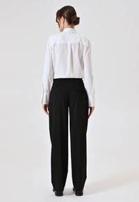 Blusa bianca a maniche lunghe con retro plissettato, abbinata a pantaloni neri a vita alta con due tasche posteriori e una linea dritta.