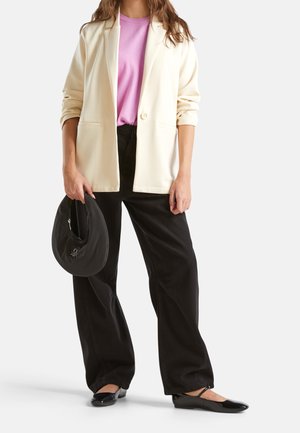 Femme portant un blazer crème, un top rose, un pantalon large noir et des ballerines noires, tenant un sac à main noir sur fond blanc.
