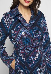 Płaszcz trench w kolorze granatowym z żywym wzorem paisley w odcieniach czerwieni, niebieskiego i białego. Posiada kołnierz, guziki oraz detal w postaci zawiązanego pasa.