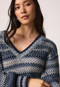 Pull à col en V en laine bleue et beige avec un motif de tricot texturé et rayé. Il possède des manches larges et des poignets côtelés.