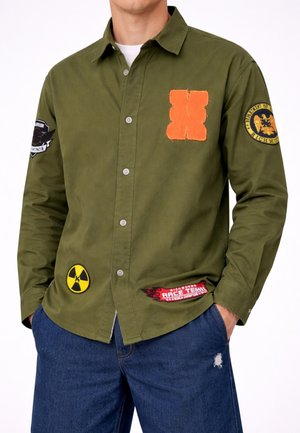 Uomo che indossa una camicia verde oliva con bottoni e toppe arancioni, gialle, nere e rosse, abbinata a jeans blu con una piccola strappatura sulla coscia sinistra.
