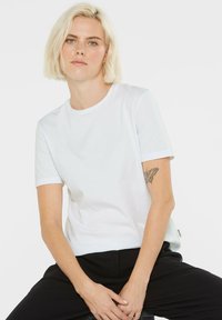 Weißes Baumwoll-T-Shirt mit kurzen Ärmeln, runden Ausschnitt und lässigem Schnitt. Verfügt über ein kleines Markenetikett an der Seite. Glatte Textur.