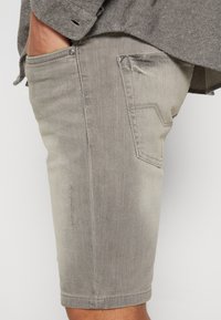 Pantaloni corti in denim grigio da uomo con una vestibilità slim, dotati di una tasca laterale e dettagli di cucitura a contrasto. Il materiale sembra morbido con una sottile trama.