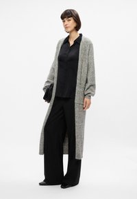 Larga chaqueta gris hecha de material suave, llevada sobre una camisa negra de botones, combinada con pantalones anchos negros y zapatos de punta.