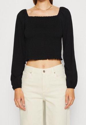 Top corto nero con scollatura quadrata, texture arricciata e maniche lunghe a sbuffo. Abbinato a pantaloni palazzo di colore chiaro.