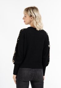 Pulover negru cu mâneci raglan, decorat cu paiete circulare aurii pe umeri. Bază și manșete striate, textură moale.