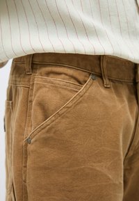 Καφέ παντελόνι κοτλέ με υφή, που διαθέτει μπροστινή τσέπη με μεταλλικό κουμπί και εμφανή ραφή κατά μήκος της μέσης.