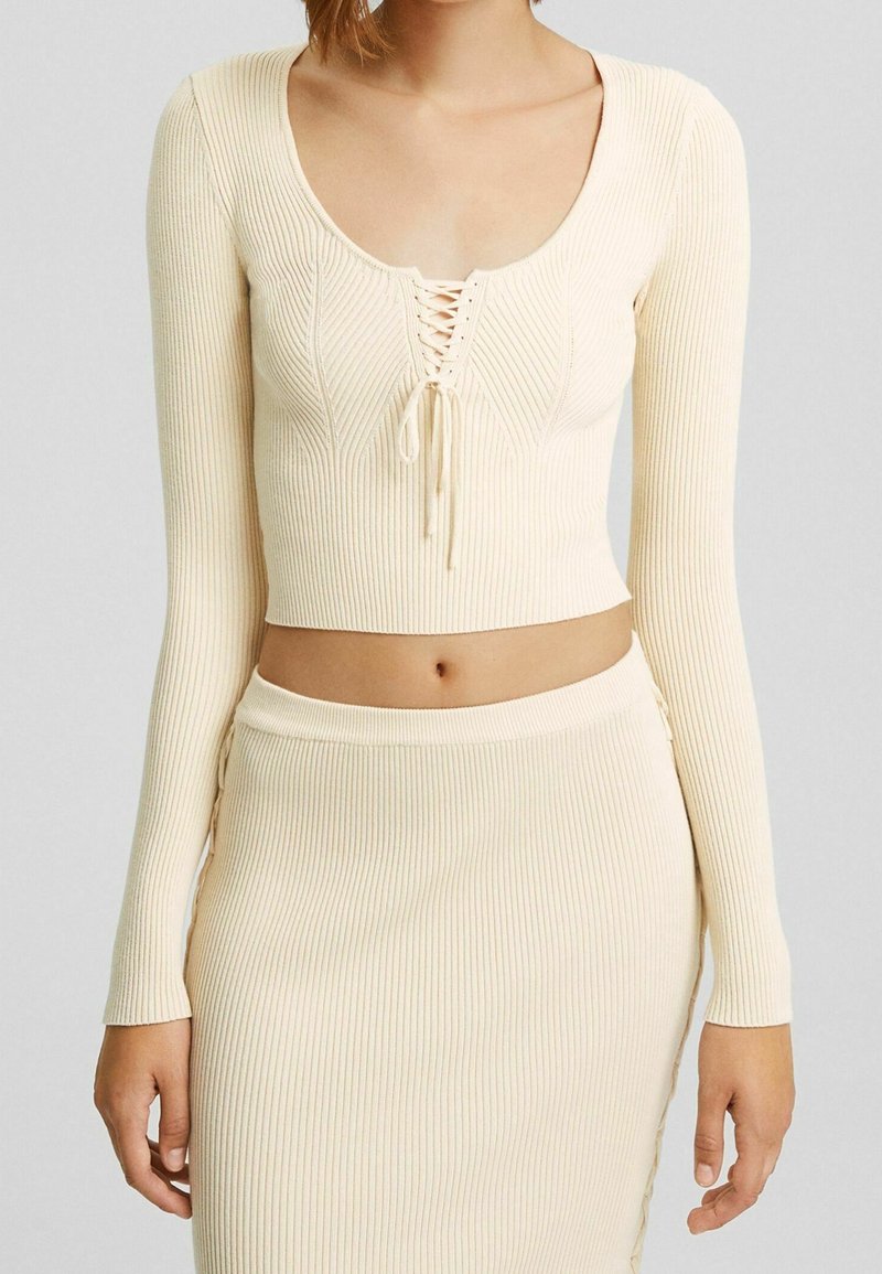 Kobieta w kremowej, prążkowanej, dzianinowej bluzce crop top z wiązaniem z przodu oraz pasującej, wysokiej spódnicy prążkowanej.