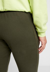 Olivgrön joggers tillverkade av mjukt, elastiskt tyg, med en slät yta och sidfickor. En lysande gulgrön sweatshirt syns ovanför.