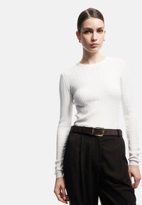 Weißes Strick-Oberteil mit langen Ärmeln, kombiniert mit dunklen Hosen und einem braunen Gürtel mit goldfarbener Schnalle. Glatte Textur und figurbetontes Design.