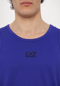 Modra brezrokavnik srajca iz mehke tkanine, z okroglim izrezom. Črn logotip EA7 Emporio Armani natisnjen na prsih.