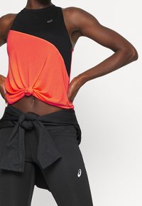 Frau mit Händen auf den Hüften, die ein ärmelloses schwarzes und oranges Oberteil trägt, das vorne gebunden ist, sowie schwarze Leggings und eine um die Taille gebundene Jacke.