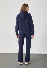 Hoodie azul marinho com cordão, corte descontraído, punhos canelados e calças combinando em tecido suave. Detalhe notável do capuz na gola.