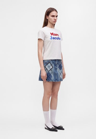 Γυναίκα στέκεται φορώντας ένα λευκό t-shirt Marc Jacobs, μπλε τζιν φούστα με μοτίβο διαμαντιού, λευκές κάλτσες και μαύρα ψηλά παπούτσια με αγκράφες.