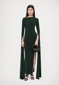 Hosszú ujjú zöld ruha oldalsó hasítékokkal, légies ujjak, karcsú sziluett és sima textúra jellemzi. Fekete kézitáskával kiegészítve.