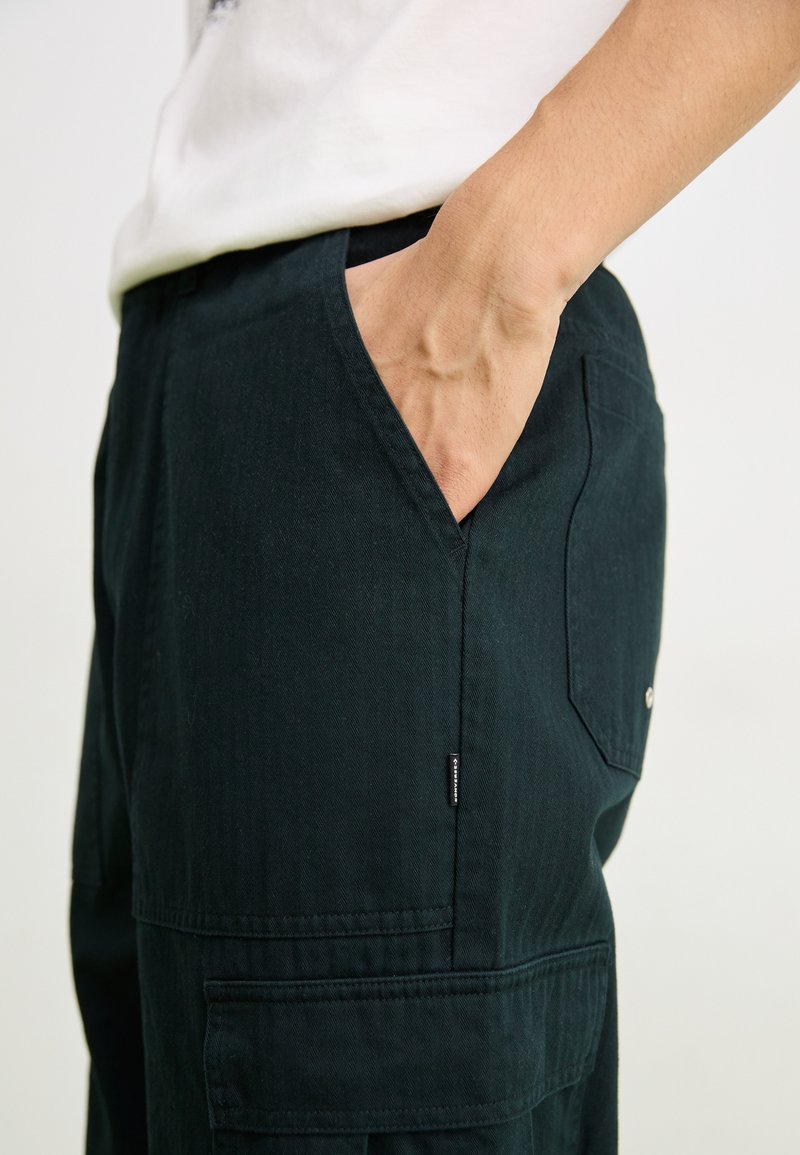 Pantalons cargo vert foncé en tissu texturé, avec des poches latérales et une étiquette de marque visible près de la couture.