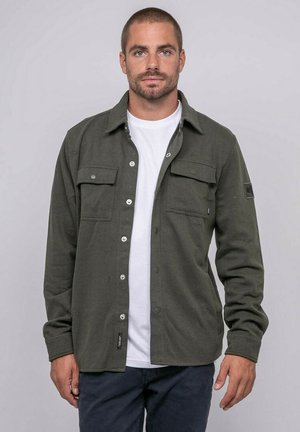 Homme portant une surchemise à boutons vert foncé par-dessus un t-shirt blanc et un pantalon sombre, debout devant un fond clair uni.