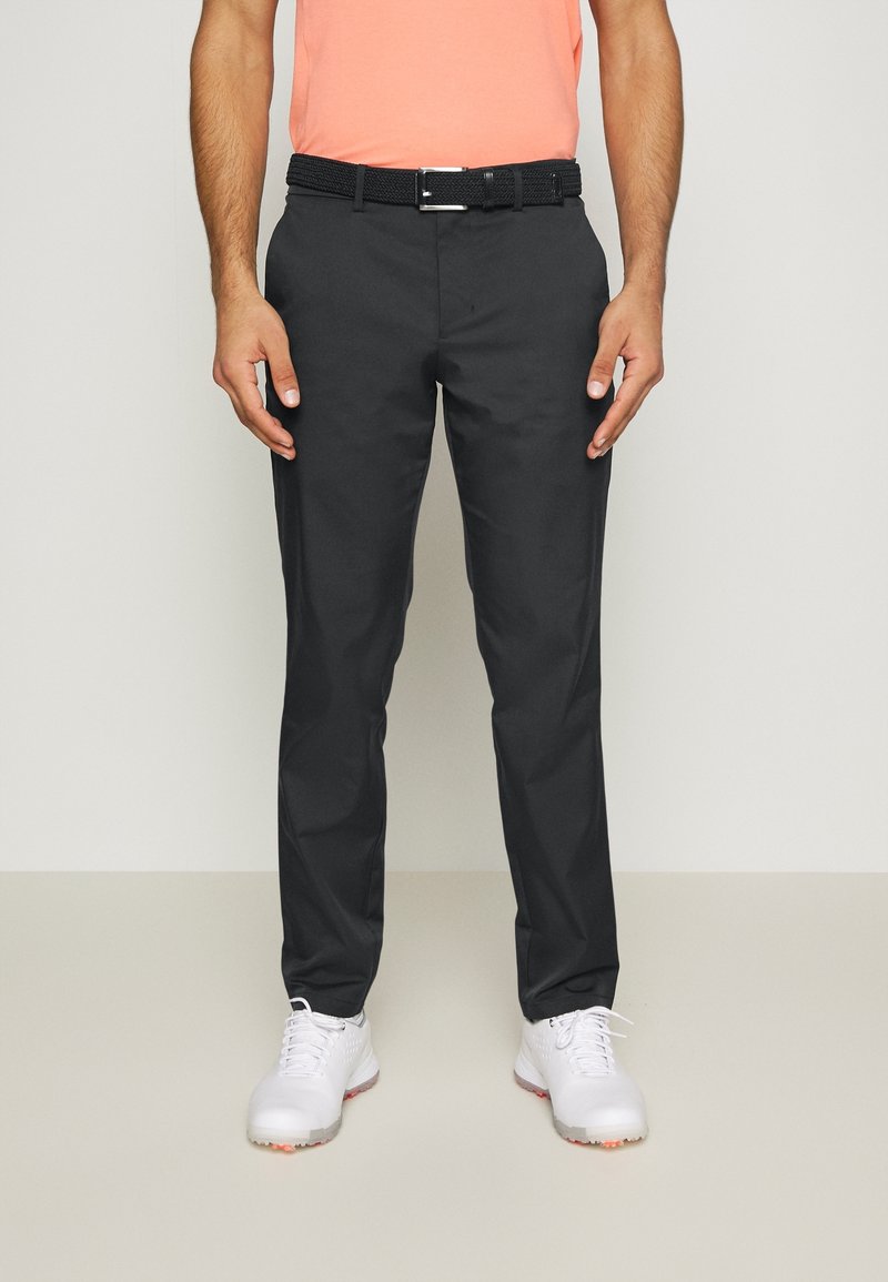 Man som bär svarta raka byxor, svart vävt bälte, korallfärgad skjorta och vita sportskor, som står mot en neutral bakgrund.