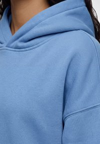 Gros plan d'une personne portant un sweat à capuche bleu clair, montrant la capuche et la zone des épaules du vêtement.