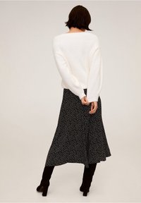 Vrouw die achteruit staat in een wit longsleeve sweater, een zwart-wit gemarmerde midi-rok en zwarte haklaarzen tegen een effen achtergrond.