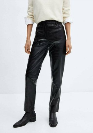 Personne portant un pantalon en cuir noir texturé, des bottines noires et un haut blanc à manches longues avec les poignets retroussés.