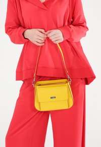 Gelbe Lederhandtasche mit rechteckiger Form und einer oberen Klappe, gehalten von einer Person in einem roten Hoodie und passenden Hosen. Silberne Hardware-Akzente.