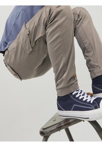 Béžové cargo kalhoty s elastickými manžetami, se bočními kapsami. Model má na sobě tmavě modré vysoké tenisky s bílými podrážkami a tkaničkami.