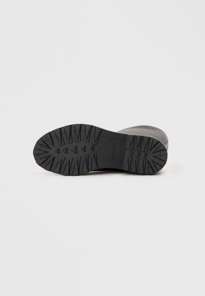 Semelle en caoutchouc noir avec un motif de texture, présentant des flèches surélevées pour l'adhérence. Partie supérieure en matériau gris avec une finition lisse.
