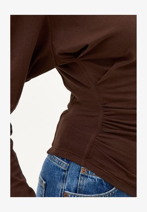 Gros plan sur une personne portant un haut marron ajusté à manches longues avec des coutures latérales froncées et un jean en denim bleu.