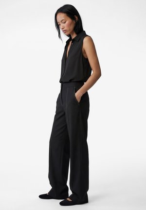 Kvinde iført sort ærmeløs bluse og løstsiddende sorte bukser, stående med hænderne i lommerne og kigger nedad mod en ensfarvet hvid baggrund.